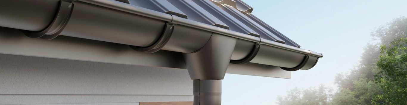 Dachrinne und Fallrohr an einem Wohngebäude mit Baum im Hintergrund.
