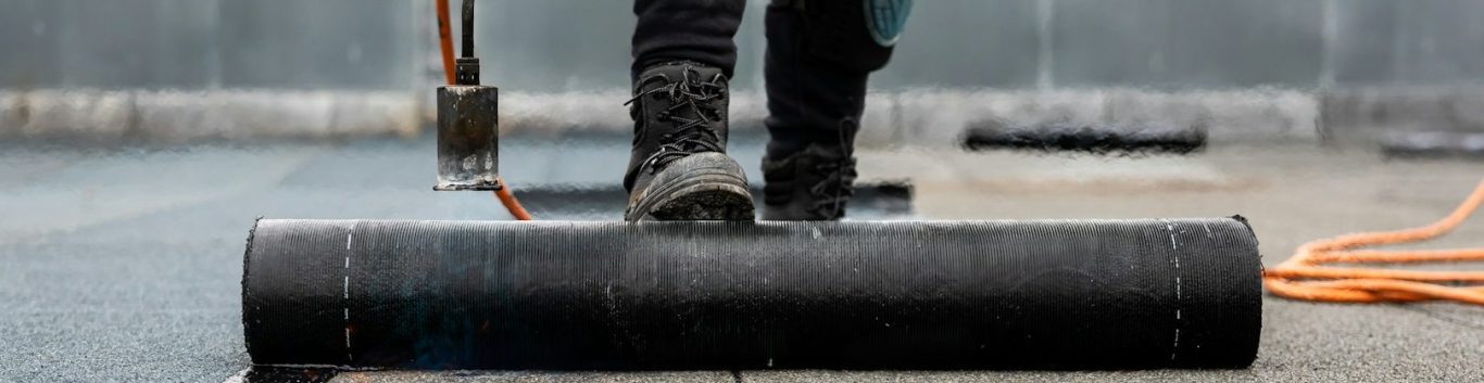 Arbeiter verlegt schwarze Bitumenbahn auf einer Fläche mit orangefarbenem Kabel.