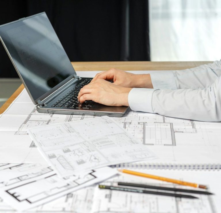 Eine Person tippt auf einem Laptop, umgeben von Bauplänen und Notizen.